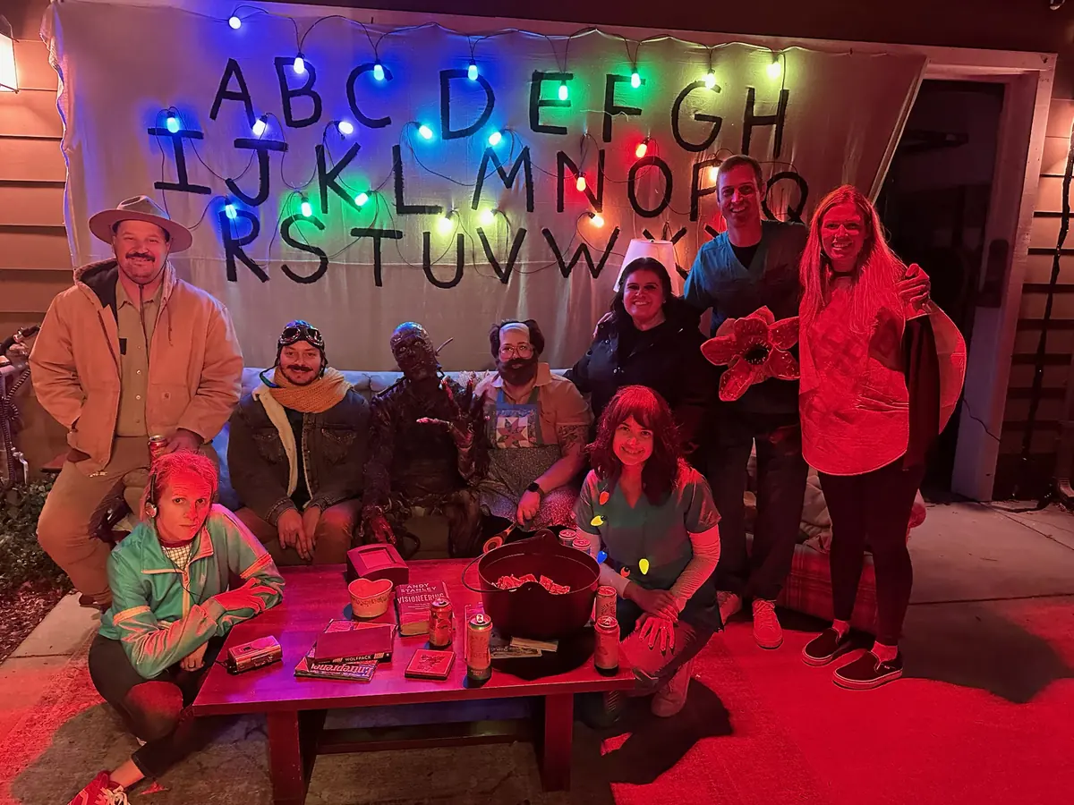 A group in costume posing in front of a hand-painted alphabet wall strung with colored Christmas lights.