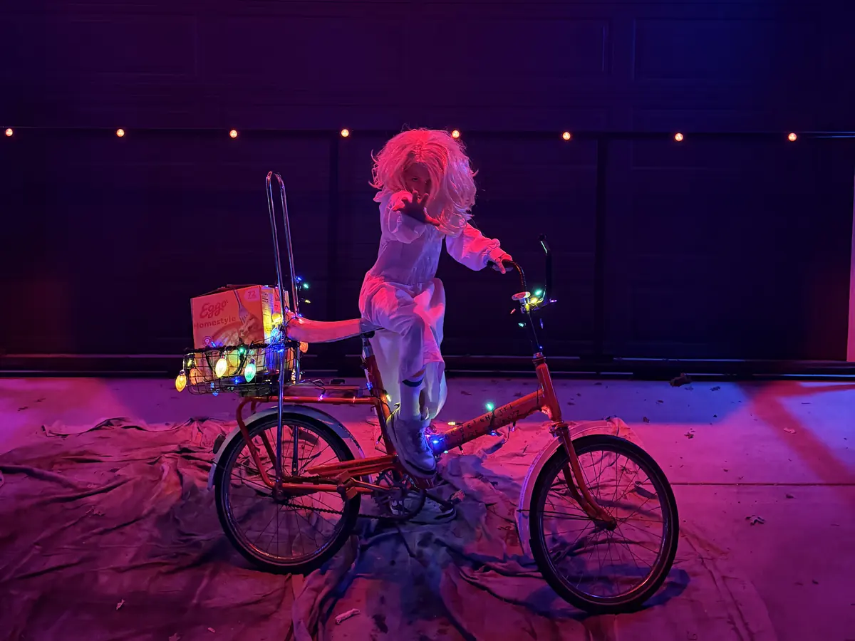 A costumed figure in a curly wig standing on a red bike with a crate on the handlebars, lit pink in front of a garage.