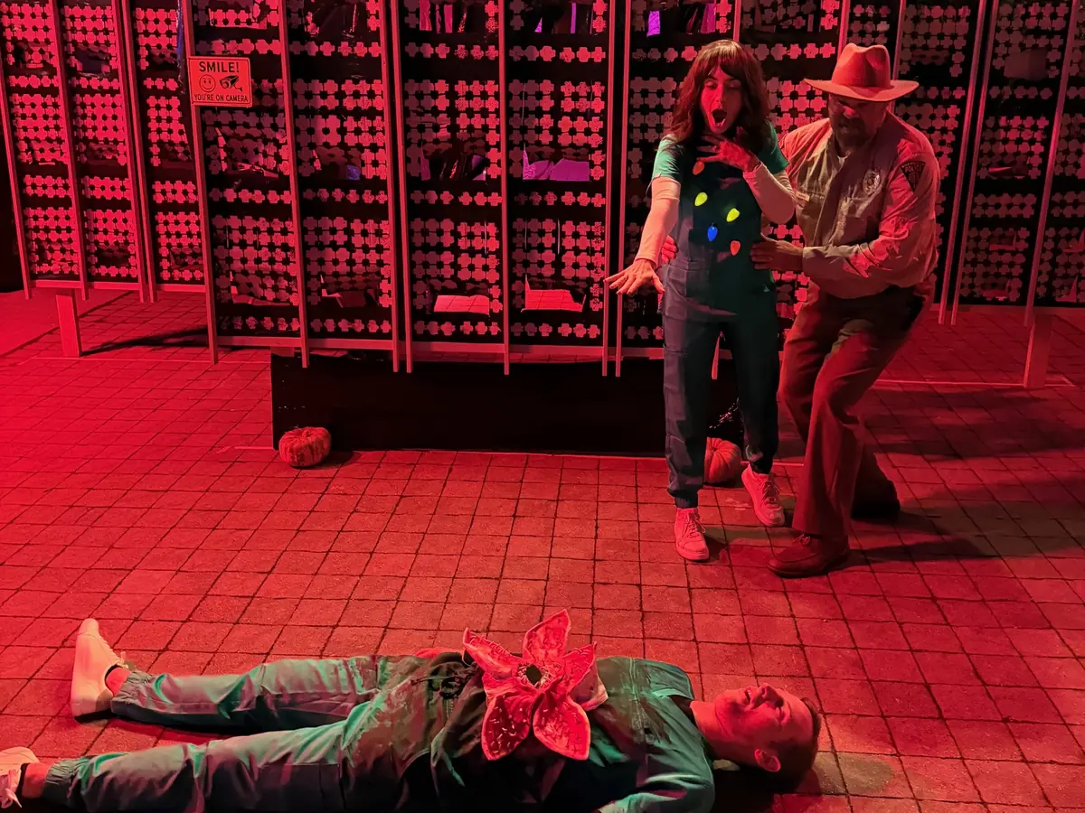 A person lying on the red-lit pavement with a Demogorgon mask while two people in costume act shocked behind them, in front of the panel wall.