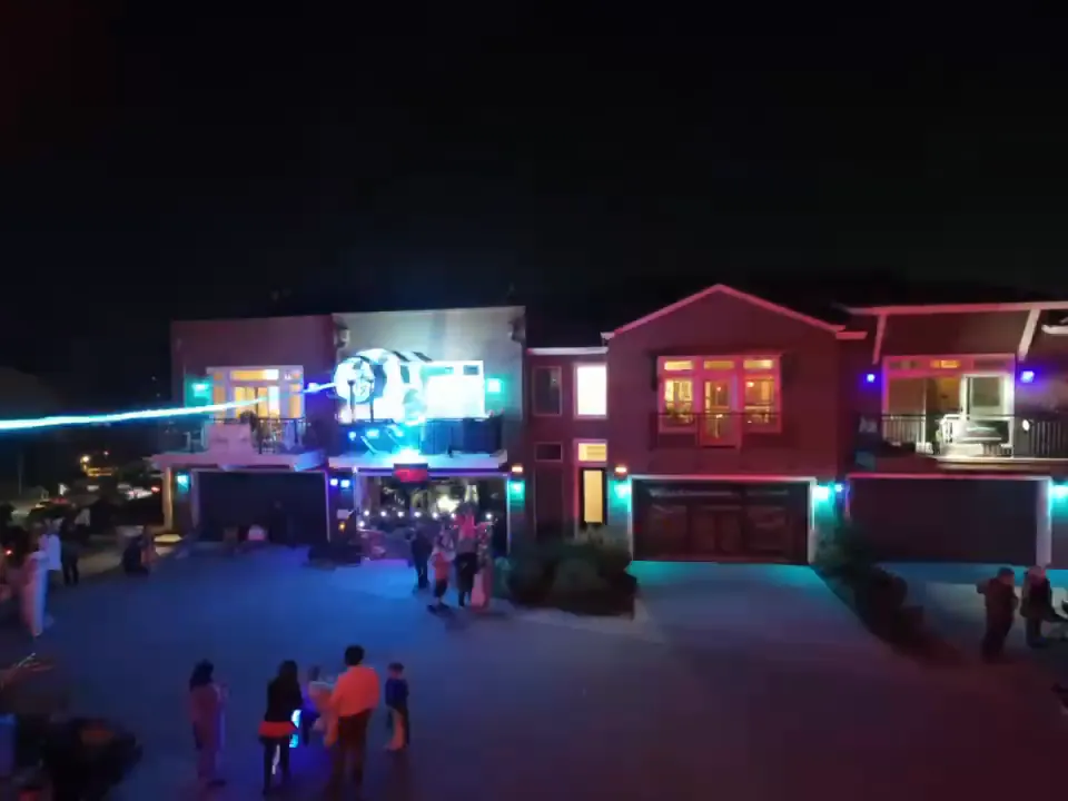 An aerial view at night: a blue beam cuts across the court from the portal while visitors watch from the street below.