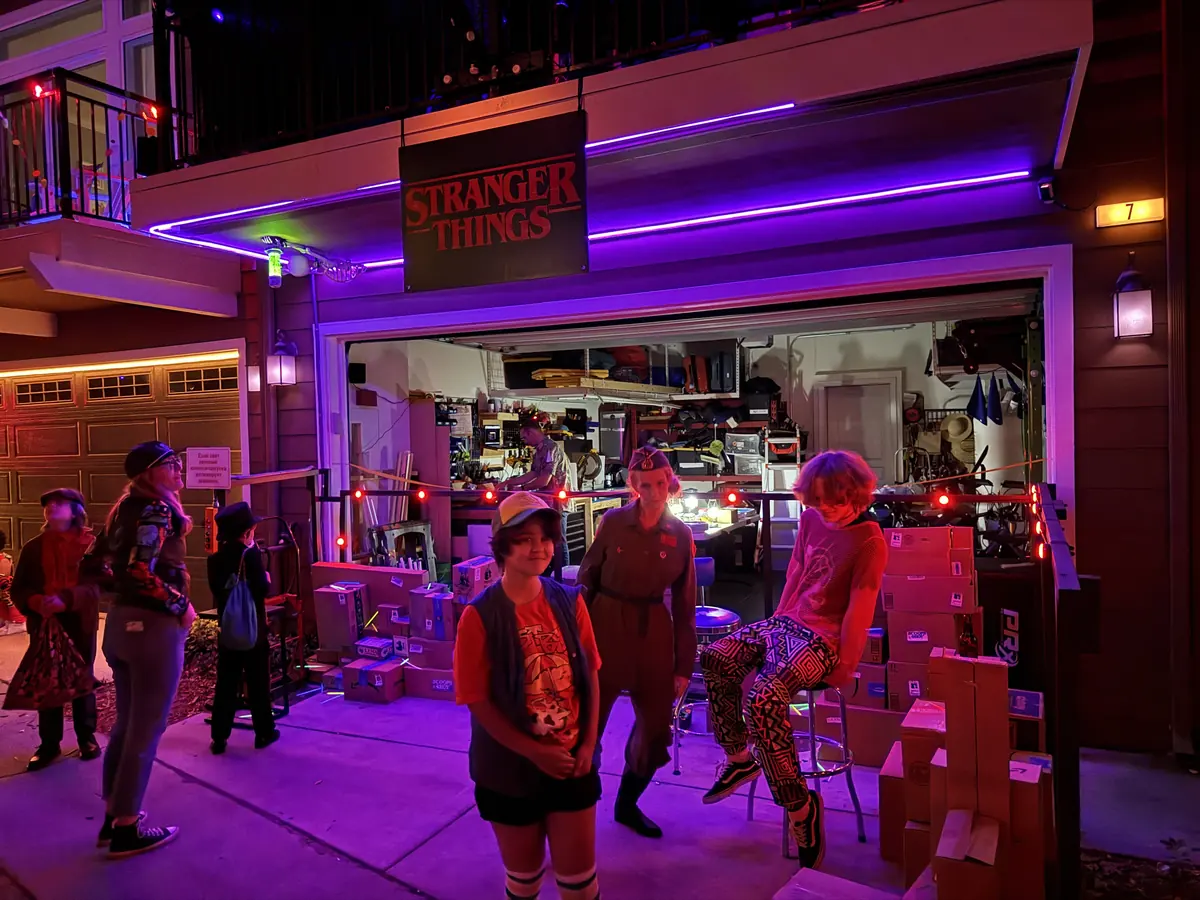 An open garage under a Stranger Things sign, lit purple, with people in eighties costumes hanging out in front.
