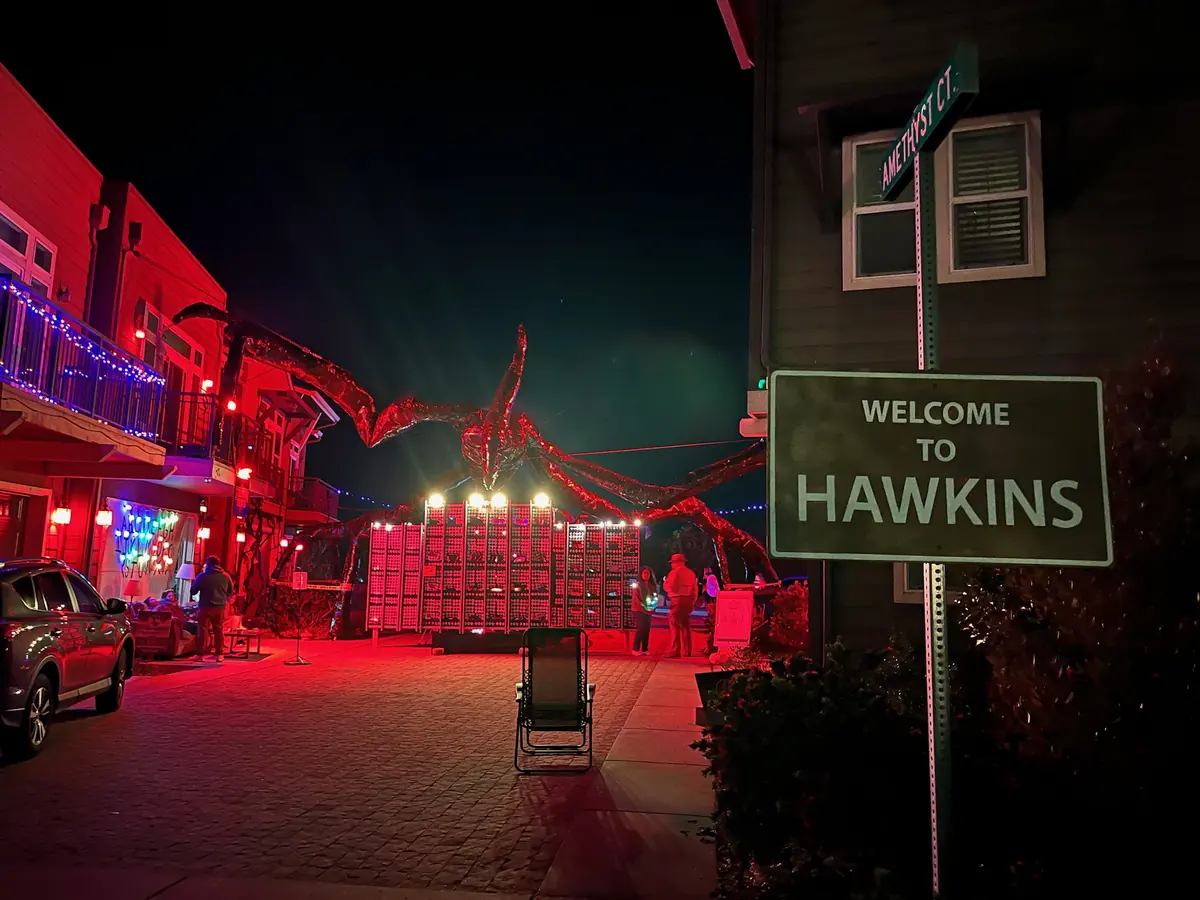 A green Welcome to Hawkins sign at the entrance to the court at night, the Mind Flayer and the light wall glowing red down the street.
