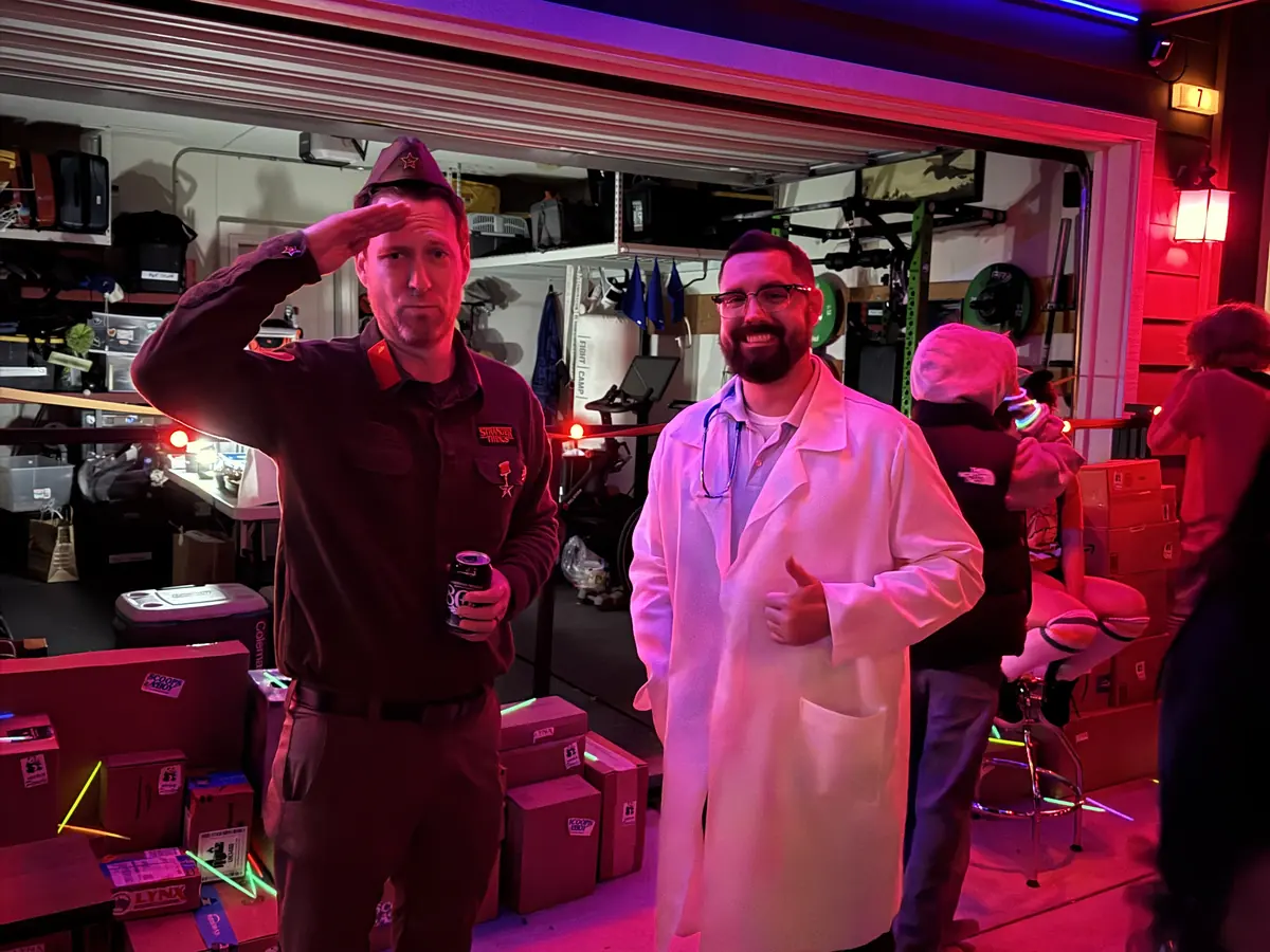 Two people in costume in an open garage, one in a police chief's uniform saluting, the other in a white lab coat giving a thumbs up.