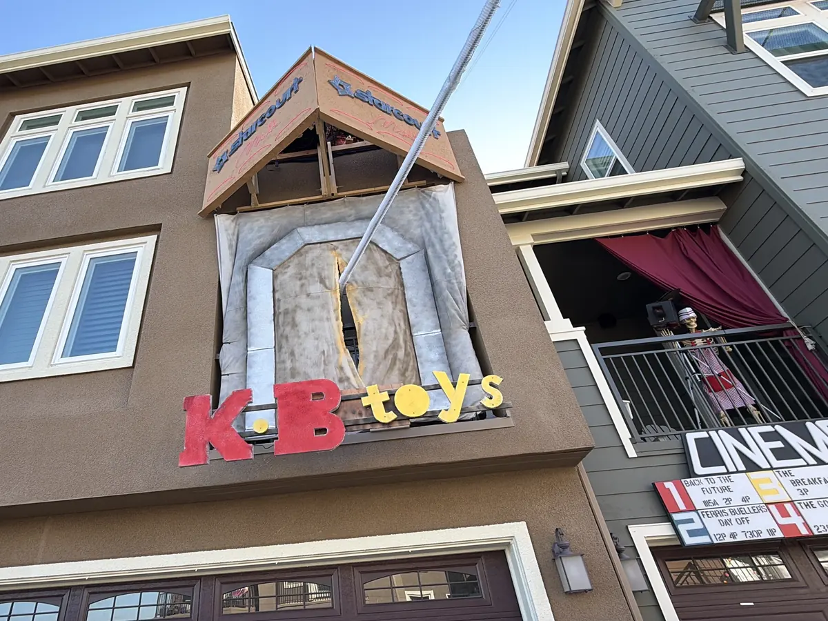 A house front dressed as a KB Toys store in daylight, with a cinema marquee on the neighboring balcony.