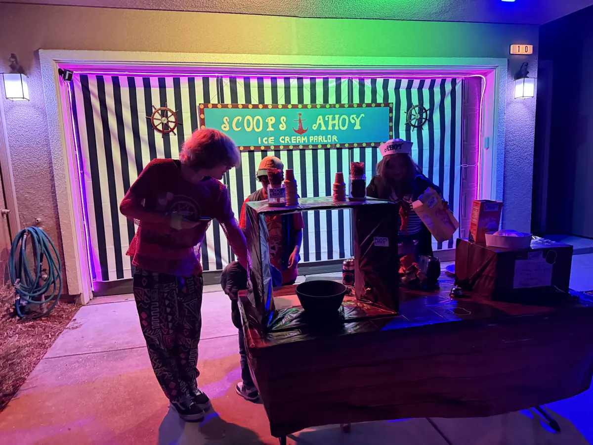 A Scoops Ahoy ice cream parlor set built into a garage, with a striped backdrop, rainbow lights and people in costume at the counter.