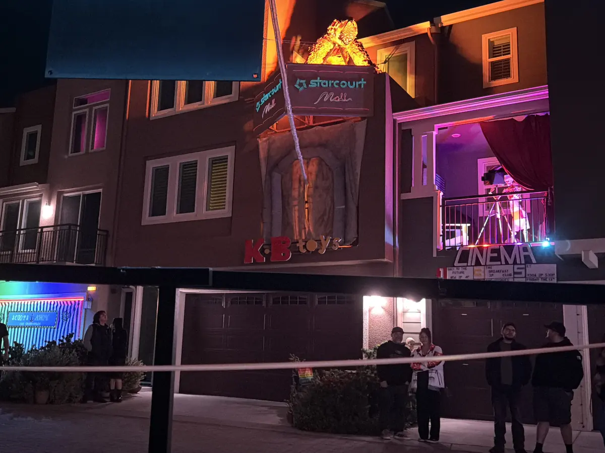 A house front dressed as Starcourt Mall at night, with a KB Toys storefront, a cinema marquee on the balcony, and visitors along the rope line.
