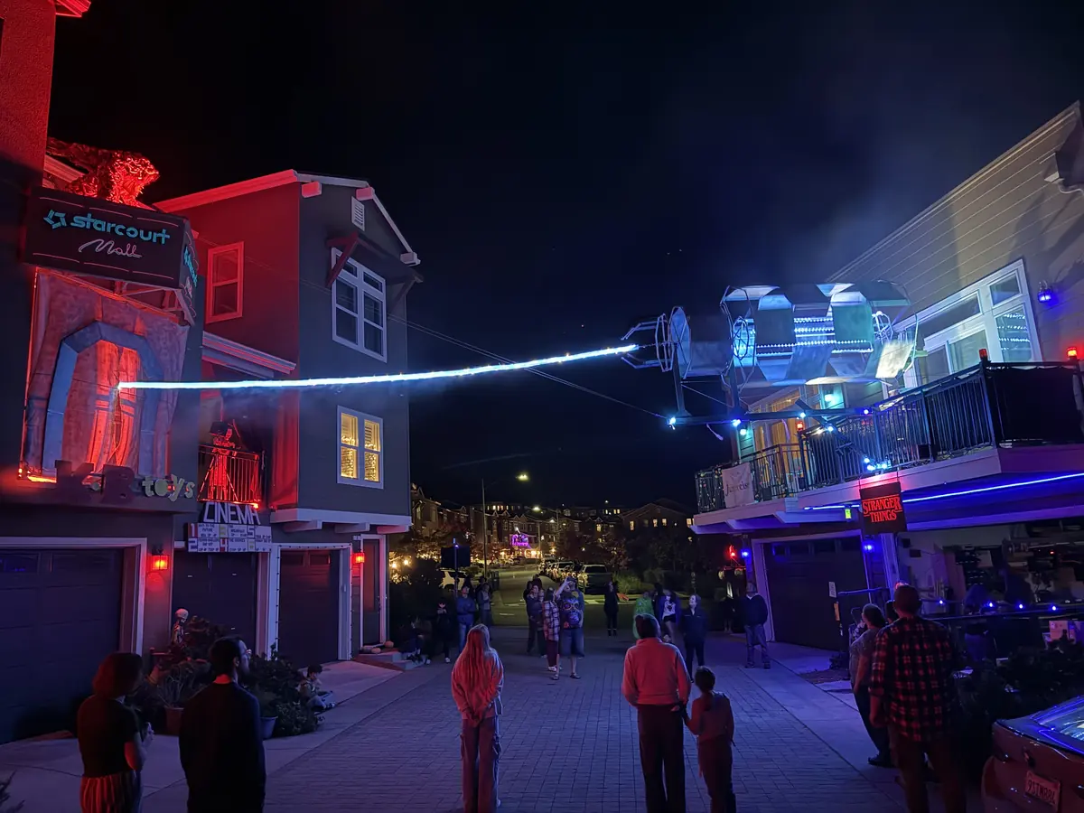 The cul-de-sac at night full of visitors, a glowing cable strung overhead to a lit rig on a balcony, the Starcourt Mall sign on the left.
