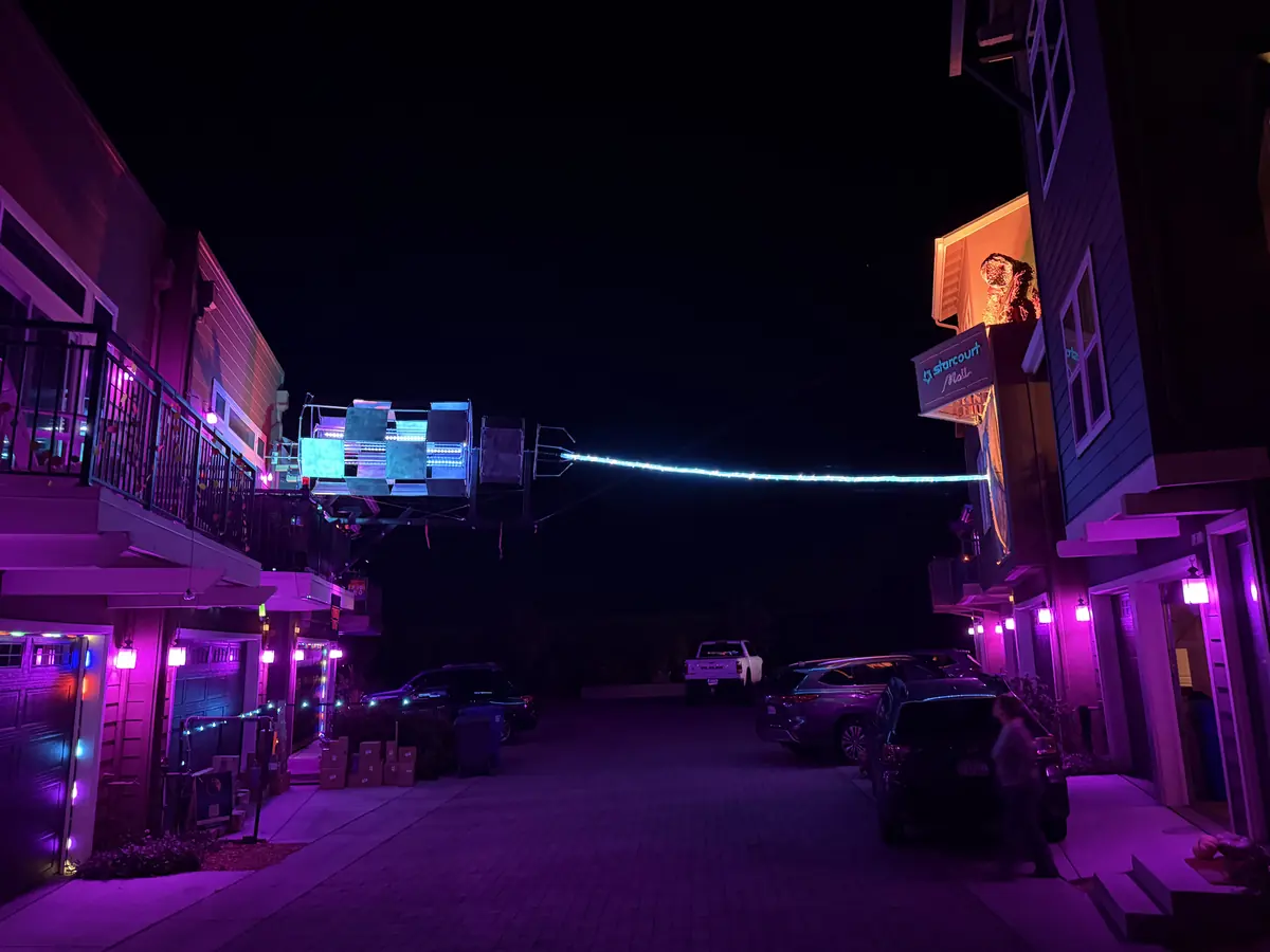 The empty cul-de-sac at night, the houses washed in purple light and a glowing cable running to the rig overhead.