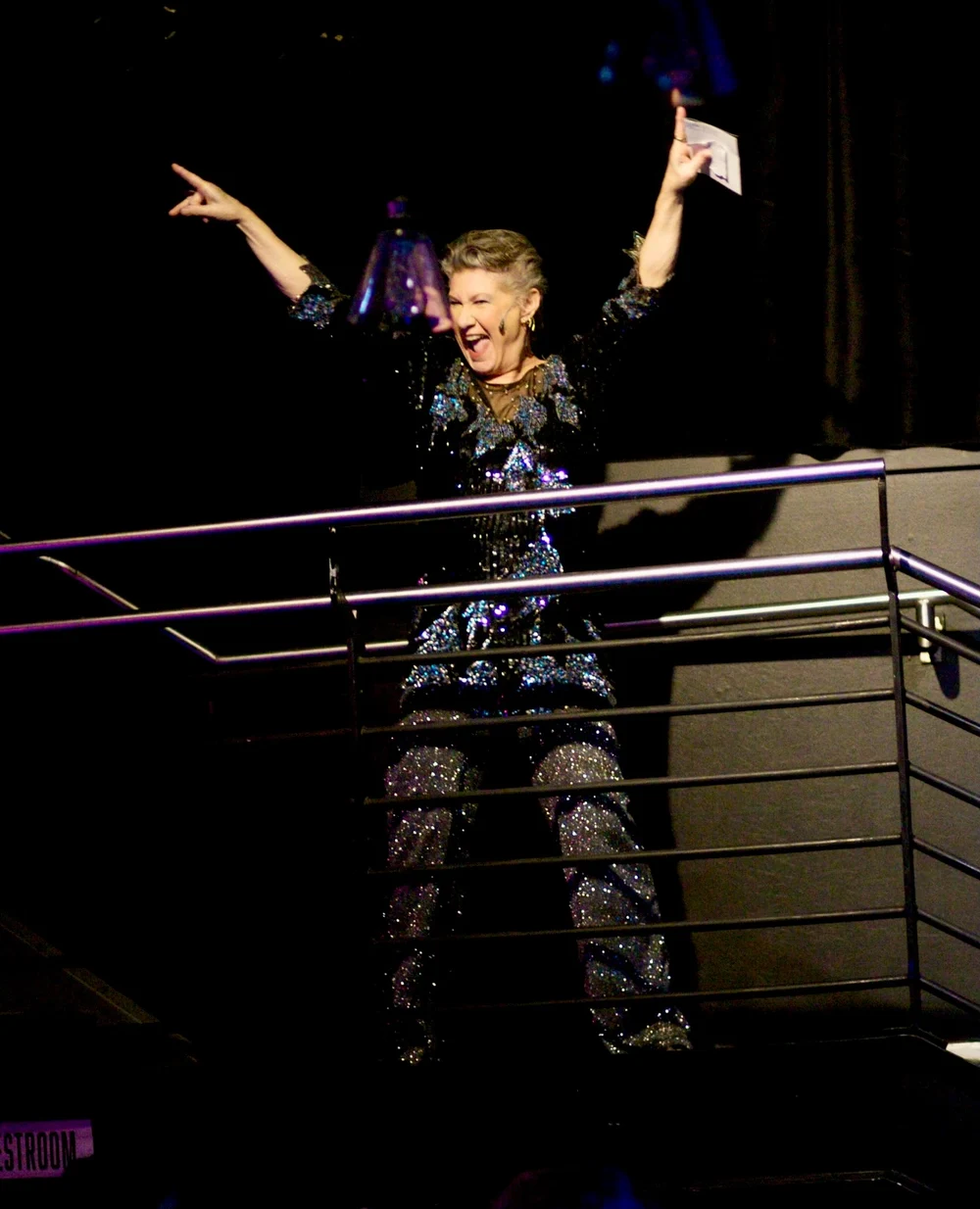 Cheryl King in a sequined jumpsuit, arms raised, on a theatre balcony