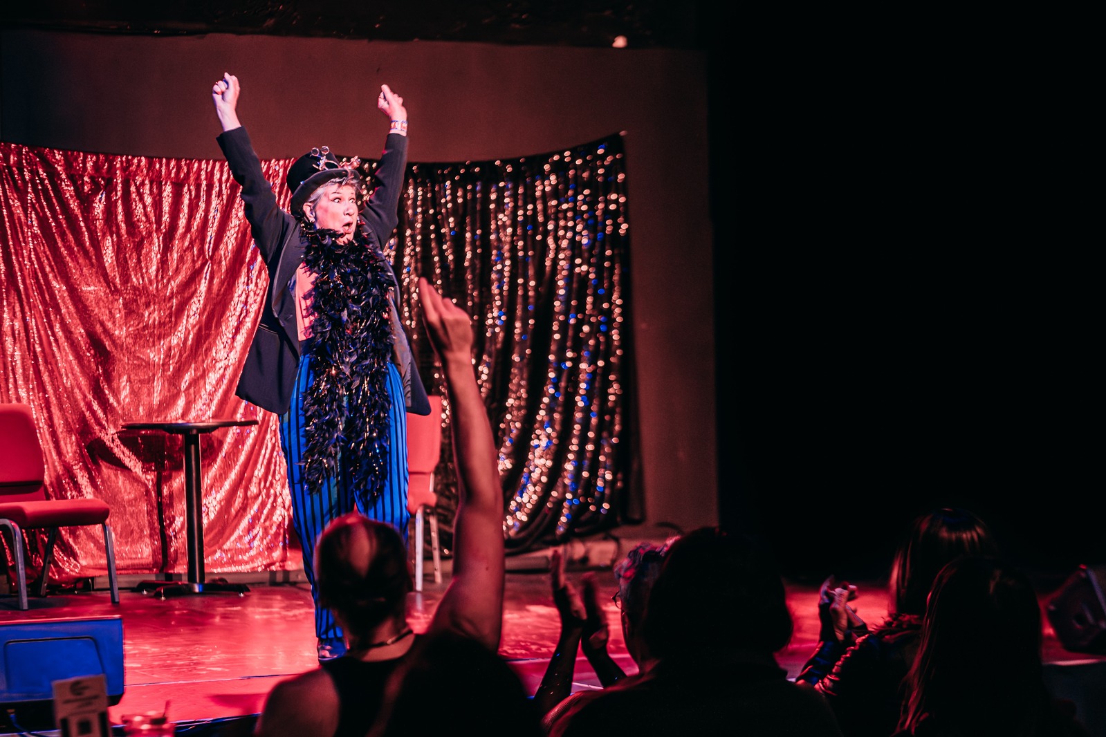 A performer in a feather boa raises both arms in front of a gold curtain as the audience cheers